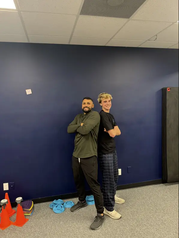 A Greiner physical therapist and a teenage athlete standing back to back with arms crossed in the clinic